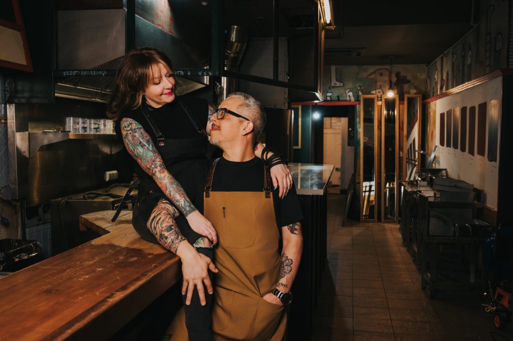 Sacred Slice Pizza owners smiling at each other in a candid moment inside their Barcelona pizzeria kitchen